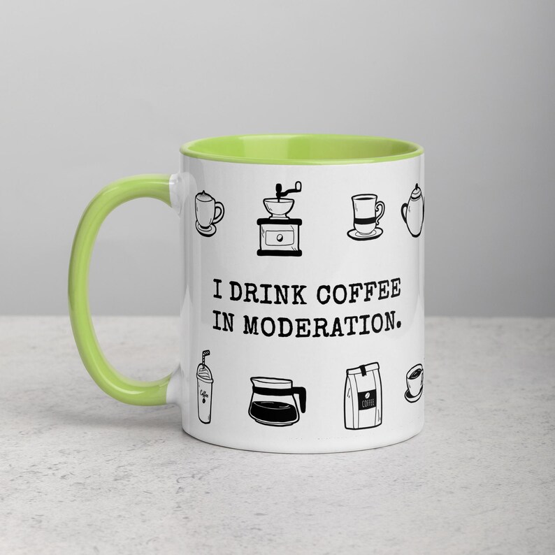 Op de afbeelding: Witte keramische koffiemok met een lichtgroen interieur en handvat. De mok is versierd met zwarte lijntekeningen van koffiegerelateerde items en de tekst "I DRINK COFFEE IN MODERATION."