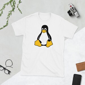 Puede incluir: Camiseta blanca con un dise&ntilde;o de ping&uuml;ino de dibujos animados. El ping&uuml;ino es negro y amarillo, sentado con los brazos a los lados. La camiseta tiene cuello redondo y es de material suave.