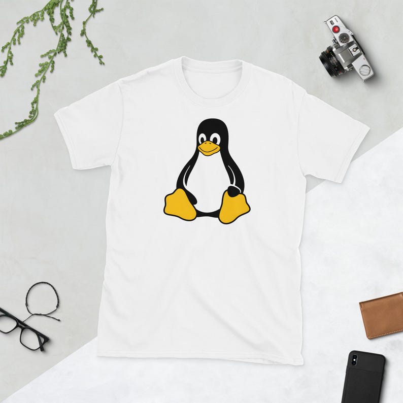 Puede incluir: Camiseta blanca con un dise&ntilde;o de ping&uuml;ino de dibujos animados. El ping&uuml;ino es negro y amarillo, sentado con los brazos a los lados. La camiseta tiene cuello redondo y es de material suave.