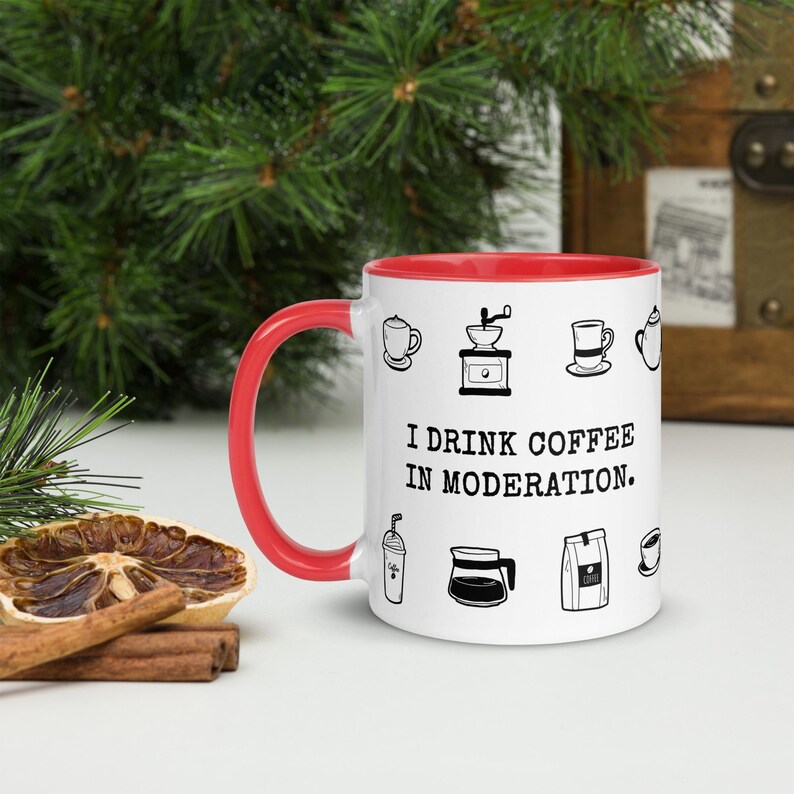 Op de afbeelding: Witte koffiemok met een rode binnenkant en handvat. De mok heeft zwarte lijntekeningen van koffiegerelateerde items en de tekst "I DRINK COFFEE IN MODERATION." De mok staat op een wit oppervlak met een gedroogde sinaasappelschijf en kaneelstokjes.