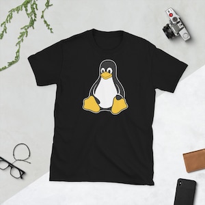 Puede incluir: Camiseta negra con un dise&ntilde;o de ping&uuml;ino de dibujos animados. El ping&uuml;ino tiene un vientre blanco, un cuerpo negro y pies amarillos. La camiseta es de cuello redondo y manga corta. El ping&uuml;ino est&aacute; sentado.
