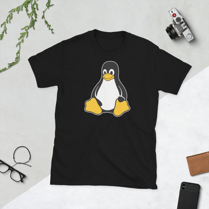 Puede incluir: Camiseta negra con un dise&ntilde;o de ping&uuml;ino de dibujos animados. El ping&uuml;ino tiene un vientre blanco, un cuerpo negro y pies amarillos. La camiseta es de cuello redondo y manga corta. El ping&uuml;ino est&aacute; sentado.