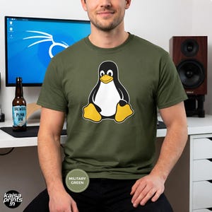 May include: Military green t-shirt featuring a cartoon penguin with a black body, white belly, and yellow feet. The shirt has a crew neck and short sleeves. The text "MILITARY GREEN" is printed on a circular label. The shirt is part of a clothing collection.