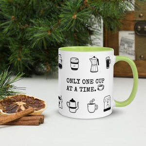 Op de afbeelding: Witte koffiemok met een groene binnenkant en handvat. De mok is versierd met zwarte lijntekeningen van koffie-gerelateerde items en de tekst "ONLY ONE CUP AT A TIME."