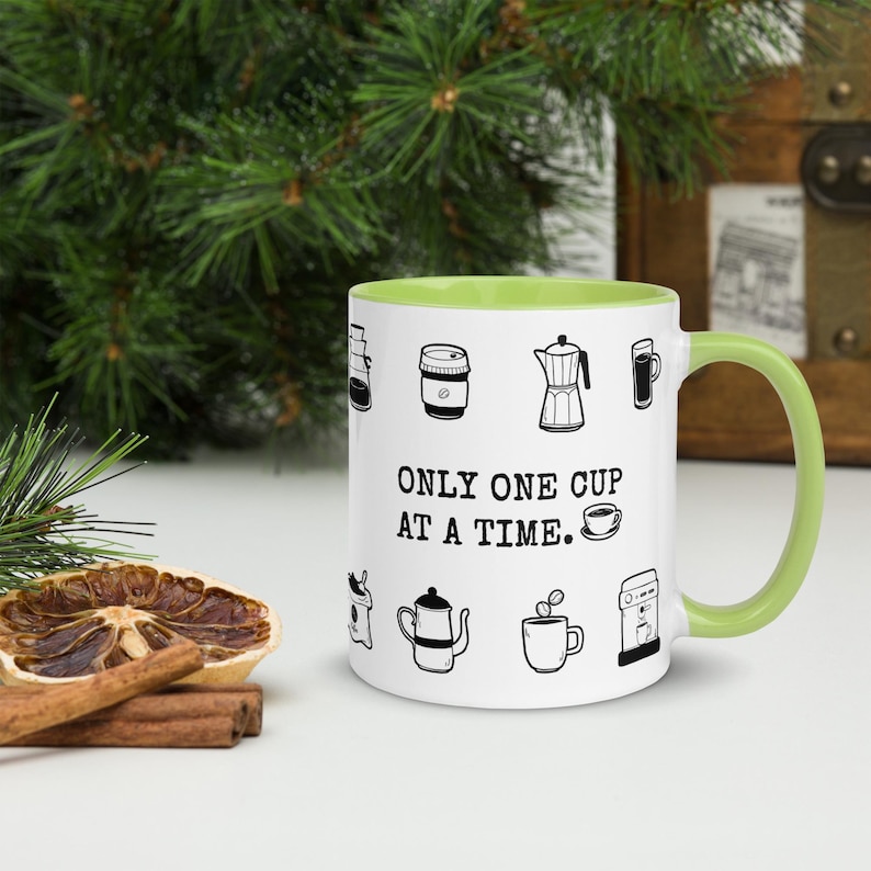 Op de afbeelding: Witte koffiemok met een groene binnenkant en handvat. De mok is versierd met zwarte lijntekeningen van koffie-gerelateerde items en de tekst "ONLY ONE CUP AT A TIME."