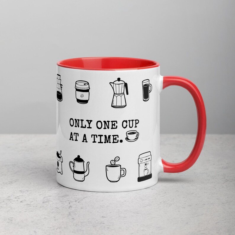 Op de afbeelding: Witte keramische koffiemok met een rode binnenkant en handvat. De mok heeft zwarte lijntekeningen van koffiegerelateerde items en de tekst "ONLY ONE CUP AT A TIME."