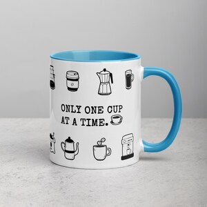 Op de afbeelding: Witte keramische koffiemok met een blauwe binnenkant en handvat. De mok heeft zwarte lijntekeningen van koffiegerelateerde items en de tekst "ONLY ONE CUP AT A TIME."