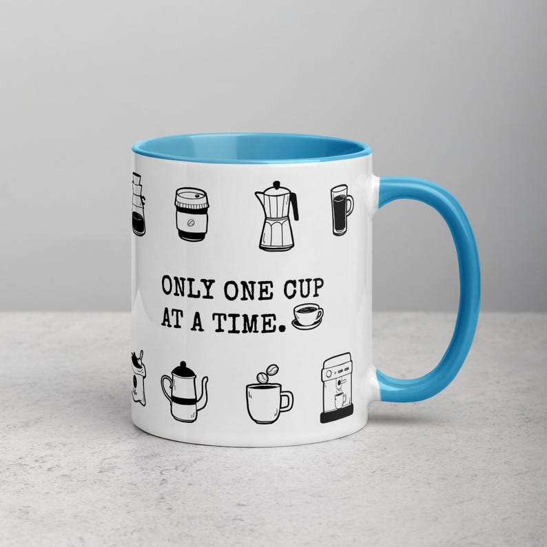 Op de afbeelding: Witte keramische koffiemok met een blauwe binnenkant en handvat. De mok heeft zwarte lijntekeningen van koffiegerelateerde items en de tekst "ONLY ONE CUP AT A TIME."
