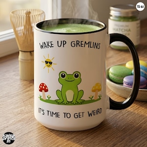 May include: A white ceramic mug with a black handle and rim, filled with green liquid. The mug features a cartoon frog, sun, and mushroom design with the text "WAKE UP GREMLINS" and "IT'S TIME TO GET WEIRD". The mug holds 15 oz.