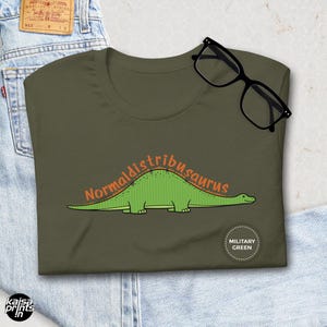 Puede incluir: Camiseta verde militar con una ilustración de dinosaurio y el texto "Normaldistribusaurus" en naranja. La camiseta está doblada sobre unos vaqueros azules, con unas gafas negras encima. Ropa informal.