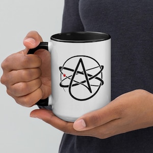 May include: White ceramic mug with a black interior and handle. The mug features a black graphic of a stylized atom with a red dot and the letter A. The mug is held by a person.