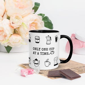 Op de afbeelding: Witte koffiemok met een zwarte binnenkant en handvat, versierd met zwarte lijntekeningen van koffiezetapparatuur. De mok heeft de tekst "ONLY ONE CUP AT A TIME". De mok staat op een oppervlak met chocolade en snoep.
