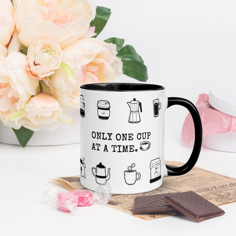 Op de afbeelding: Witte koffiemok met een zwarte binnenkant en handvat, versierd met zwarte lijntekeningen van koffiezetapparatuur. De mok heeft de tekst "ONLY ONE CUP AT A TIME". De mok staat op een oppervlak met chocolade en snoep.