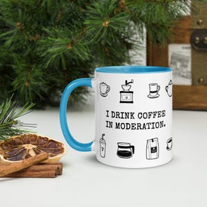 Op de afbeelding: Witte koffiemok met een blauw handvat en binnenkant, versierd met zwarte lijntekeningen met koffiethema en de tekst "I DRINK COFFEE IN MODERATION." De mok staat tegen een achtergrond van groen en een houten kist.