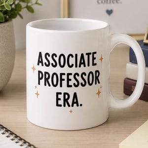 May include: A white ceramic mug featuring the black text "ASSOCIATE PROFESSOR ERA." printed on the front, accented by small orange star icons. The mug has a standard handle and a smooth finish, positioned upright on a flat surface.