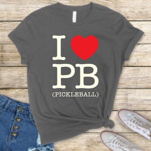 May include: A grey t-shirt with the text "I love PB (Pickleball)" in white and a red heart.