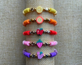 May include: Five colorful bracelets with different fruit charms. The bracelets are stacked on a beige fabric background. The charms include a lemon, orange, apple, strawberry, and grapes.