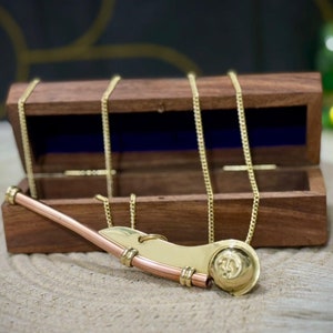 Puede incluir: Un collar con un silbato dorado y una caja de madera. El silbato es de diseño náutico tradicional con acabado en latón. La caja es de madera oscura y tiene un forro de terciopelo azul.