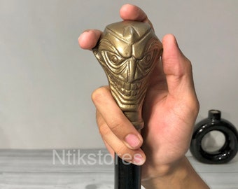 Könnte beinhalten: Ein Gehstock mit einem goldfarbenen Joker-Gesichtsdesign am Griff. Der Griff ist mit einem bedrohlichen Grinsen und scharfen Zähnen verziert. Der Stock ist schwarz und hat eine glatte Oberfläche.