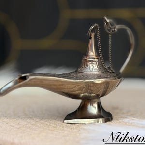 K&ouml;nnte beinhalten: Eine silberne Dschinnenlampe mit filigranen Details und einer Kette, die vom Ausguss h&auml;ngt. Die Lampe steht auf einer hellbraunen Oberfl&auml;che.