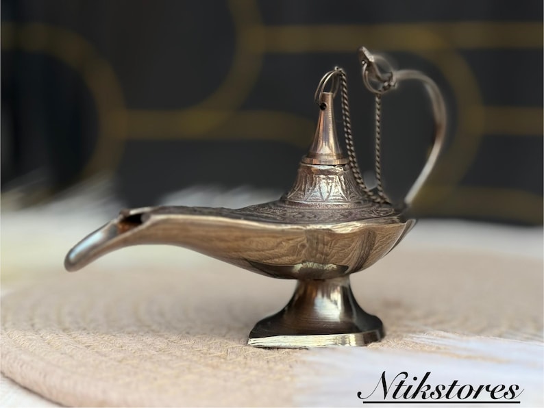 K&ouml;nnte beinhalten: Eine silberne Dschinnenlampe mit filigranen Details und einer Kette, die vom Ausguss h&auml;ngt. Die Lampe steht auf einer hellbraunen Oberfl&auml;che.