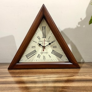 May include: A triangular wooden clock with a white face and black hands. The clock face has Roman numerals and the text "James J. Henderson Est 1823 Oxford".