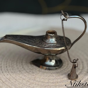 K&ouml;nnte beinhalten: Eine silberne Dschinnenlampe mit filigranen Details und einer Kette, die an einem kleinen, kegelf&ouml;rmigen Objekt aus Silber befestigt ist. Die Lampe steht auf einer gewebten Oberfl&auml;che.