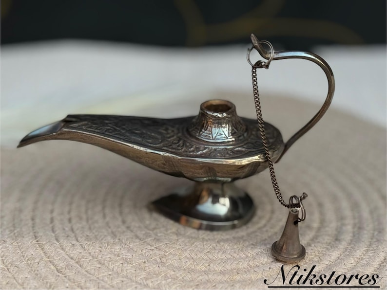 K&ouml;nnte beinhalten: Eine silberne Dschinnenlampe mit filigranen Details und einer Kette, die an einem kleinen, kegelf&ouml;rmigen Objekt aus Silber befestigt ist. Die Lampe steht auf einer gewebten Oberfl&auml;che.