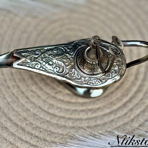 K&ouml;nnte beinhalten: Eine silberne Dschinnenlampe mit aufwendigen floralen Mustern und einem abnehmbaren Deckel. Die Lampe hat einen gebogenen Ausguss und einen Griff.