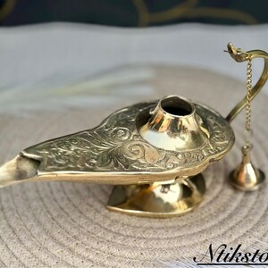 K&ouml;nnte beinhalten: Eine goldfarbene Dschinnenlampe mit aufwendigen floralen Mustern. Die Lampe hat einen Ausguss und eine kleine Kette mit einem dekorativen Quasten.