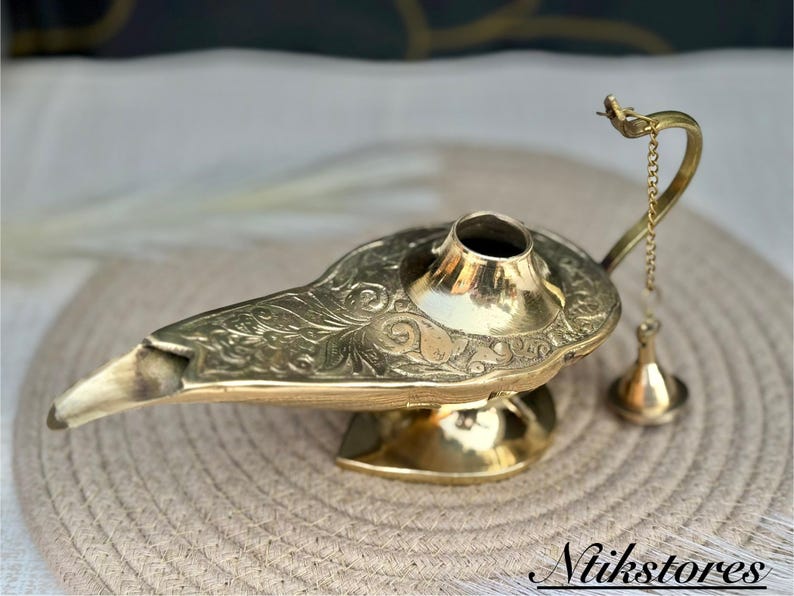 K&ouml;nnte beinhalten: Eine goldfarbene Dschinnenlampe mit aufwendigen floralen Mustern. Die Lampe hat einen Ausguss und eine kleine Kette mit einem dekorativen Quasten.