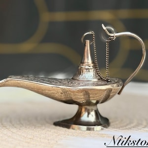 K&ouml;nnte beinhalten: Eine silberne Dschinnenlampe mit filigranen Details und einer Kette, die vom Ausguss h&auml;ngt. Die Lampe steht auf einer wei&szlig;en Oberfl&auml;che.