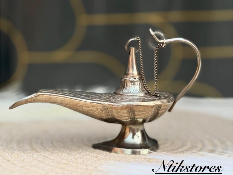 K&ouml;nnte beinhalten: Eine silberne Dschinnenlampe mit filigranen Details und einer Kette, die vom Ausguss h&auml;ngt. Die Lampe steht auf einer wei&szlig;en Oberfl&auml;che.