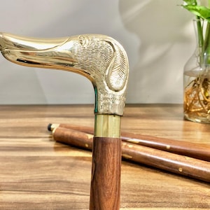 May include: A wooden walking stick with a brass dog head handle. The handle is detailed with a textured design. The stick is brown and the handle is gold.