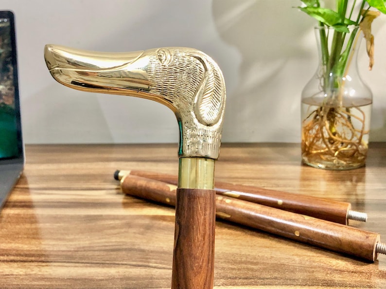 May include: A wooden walking stick with a brass dog head handle. The handle is detailed with a textured design. The stick is brown and the handle is gold.
