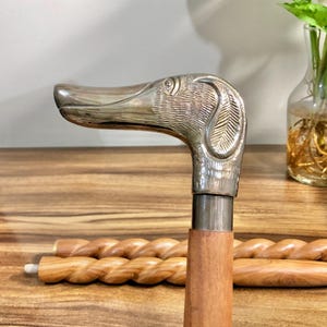 May include: A silver dog head cane handle with a wooden shaft. The cane is resting on a wooden surface.