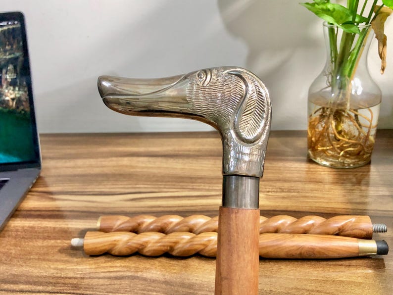May include: A silver dog head cane handle with a wooden shaft. The cane is resting on a wooden surface.