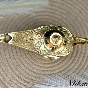 K&ouml;nnte beinhalten: Eine goldene Dschinnenlampe mit aufwendigen floralen Mustern. Die Lampe hat eine runde Oberseite und einen Ausguss an der Seite. Die Lampe steht auf einer gewebten Matte.