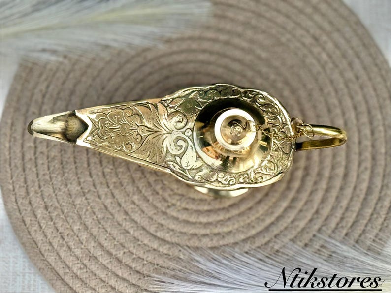 K&ouml;nnte beinhalten: Eine goldene Dschinnenlampe mit aufwendigen floralen Mustern. Die Lampe hat eine runde Oberseite und einen Ausguss an der Seite. Die Lampe steht auf einer gewebten Matte.