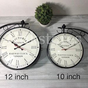 Könnte beinhalten: Zwei doppelseitige Uhren aus schwarzem Metall mit weißen Zifferblättern und schwarzen römischen Ziffern. Die größere Uhr hat einen Durchmesser von 30,5 cm, die kleinere einen Durchmesser von 25,4 cm. Beide Uhren haben schwarze Zeiger und rote Sekundenzeiger. Die Uhren zeigen "Victoria Station Railway Clock London" an.