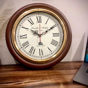 May include: A brown wooden wall clock with a brass rim and Roman numeral markings. The clock face is white with black lettering and a gold ship logo. The clock face reads "Franklin & Murphy Est. 1968 Oxford".