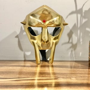 May include: A gold-colored mask with a diamond-shaped red jewel on the forehead. The mask has a sharp, angular design with a pointed chin and two horns on the sides.