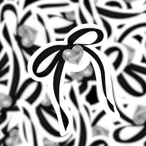 May include: A black ribbon tied into a bow with a silver heart charm that says "Priscilla" hanging from the center.