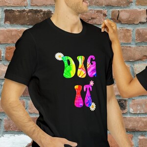 Può includere: Due magliette nere con il testo "DIG IT" in lettere tie-dye arcobaleno e margherite bianche. Le magliette sono indossate da due persone in piedi davanti a un muro di mattoni.