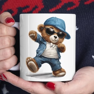 Può includere: Tazza in ceramica bianca con un'illustrazione di un orso marrone che indossa occhiali da sole, un cappello blu e una giacca di jeans blu. L'orso sta ballando.