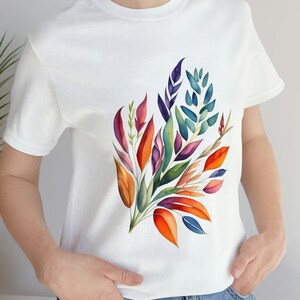 Può includere: Una t-shirt bianca con un design floreale colorato. Il design presenta un bouquet di foglie e fiori in tonalità di arancione, rosso, viola, blu e verde.