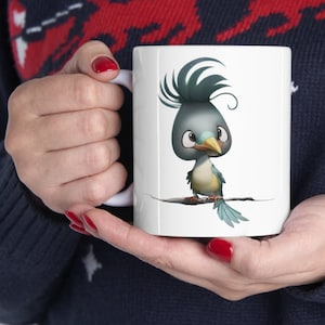 May include: White ceramic mug featuring a cartoon bird with a blue crest and teal accents. The bird is perched on a crack design. The mug is held by a person wearing a navy blue sweater with a red and white reindeer pattern.
