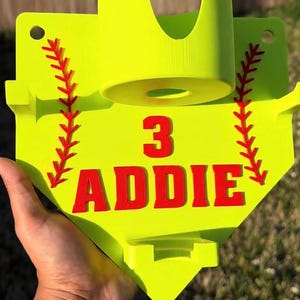 Könnte beinhalten: Eine neongelbe Wandhalterung in Form einer Homeplate mit Getränkehalter und zwei seitlichen Haken. Das Design zeigt rote, baseballartige Nahtmuster an den Seiten sowie den roten Schriftzug "3" und "ADDIE" in der Mitte des Objekts.