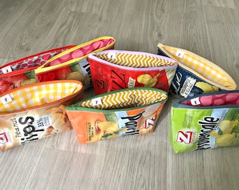 以下が含まれることがあります： 再利用されたポテトチップスの袋から作られた、カラフルなジッパー付きポーチが7つあります。ポーチには、赤、黄、緑、青、白など、さまざまなパターンと色が使われています。ポーチはすべて閉じており、木製の表面に置かれています。