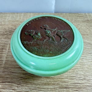 Puede incluir: Un recipiente redondo verde menta con una tapa de color bronce. La tapa presenta un relieve de tres perros en un campo, con el texto "POTTER & MOORE". El recipiente tiene un diseño liso y redondeado.